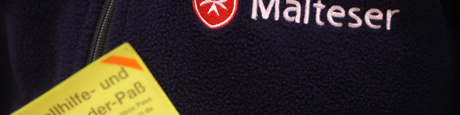 Ein gelber Blutspendepass wird vor einer blauen Malteser-Jacke gehalten.