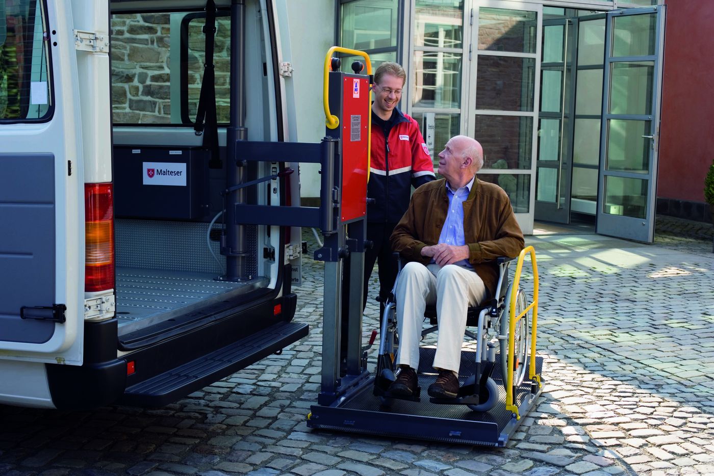 Ein Fahrstuhlfahrer steht auf einer Plattform, mit der ein Helfer der Malteser ihn in ein Fahrstuhltransportwagen rollen kann.