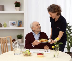 Menüservice Essen auf Rädern Malteser Lohne Eine jüngere Dame gibt einem älteren Herren, der an einem Tisch sitzt, einen Teller mit dem Mittagessen.