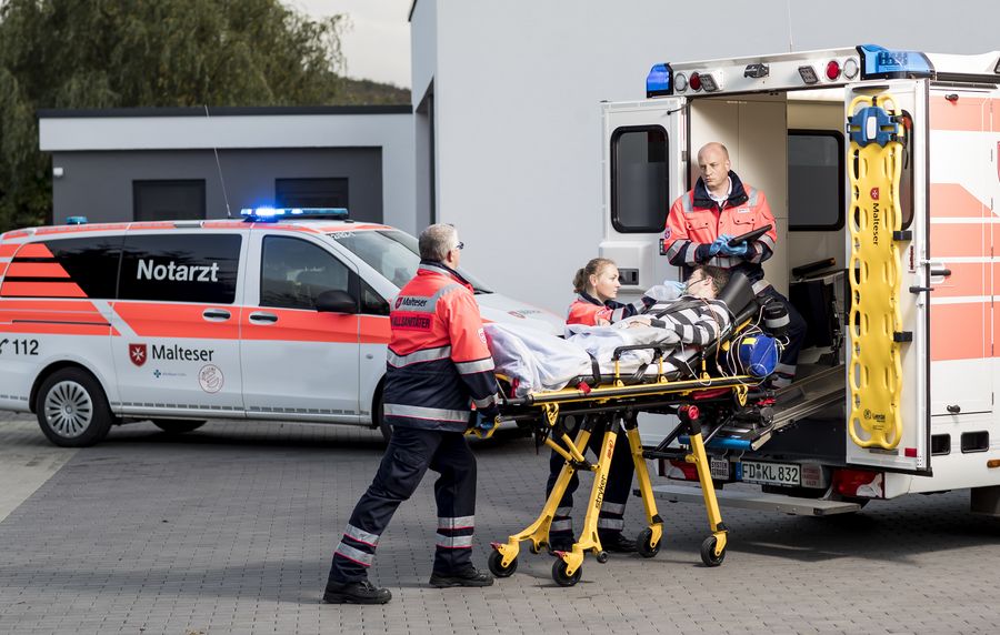 Zwei Helfer der Malteser schieben eine Krankentrage in einen Rettungswagen. Im Hintergrund ist ein weiteres Malteser-Fahrzeug zu sehen.