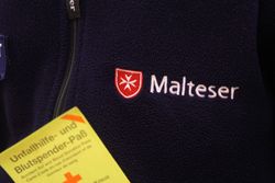 Ein gelber Blutspendepass wird vor einer blauen Jacke der Malteser gehalten.